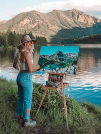 a woman painting in front of a lake with mountains in the background