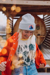 a woman wearing a beanie and jeans standing in front of a cabin